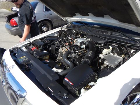 2003 Ford Crown Victoria - Close view of a white sedan's open engine bay showing a cold-air intake, with a man and a gray Crown Victoria visible in the background.