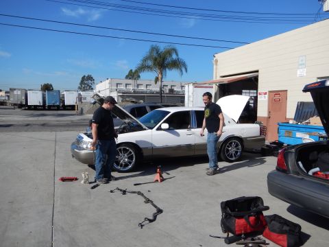 2003 Ford Crown Victoria - Two men stand beside a white Lincoln Town Car with its hood raised and suspension parts laid out on the pavement in a sunny lot.