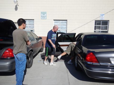 2003 Ford Crown Victoria - group of men work around the open rear door of a dark Lincoln sedan parked next to a gray Crown Victoria in an industrial lot.