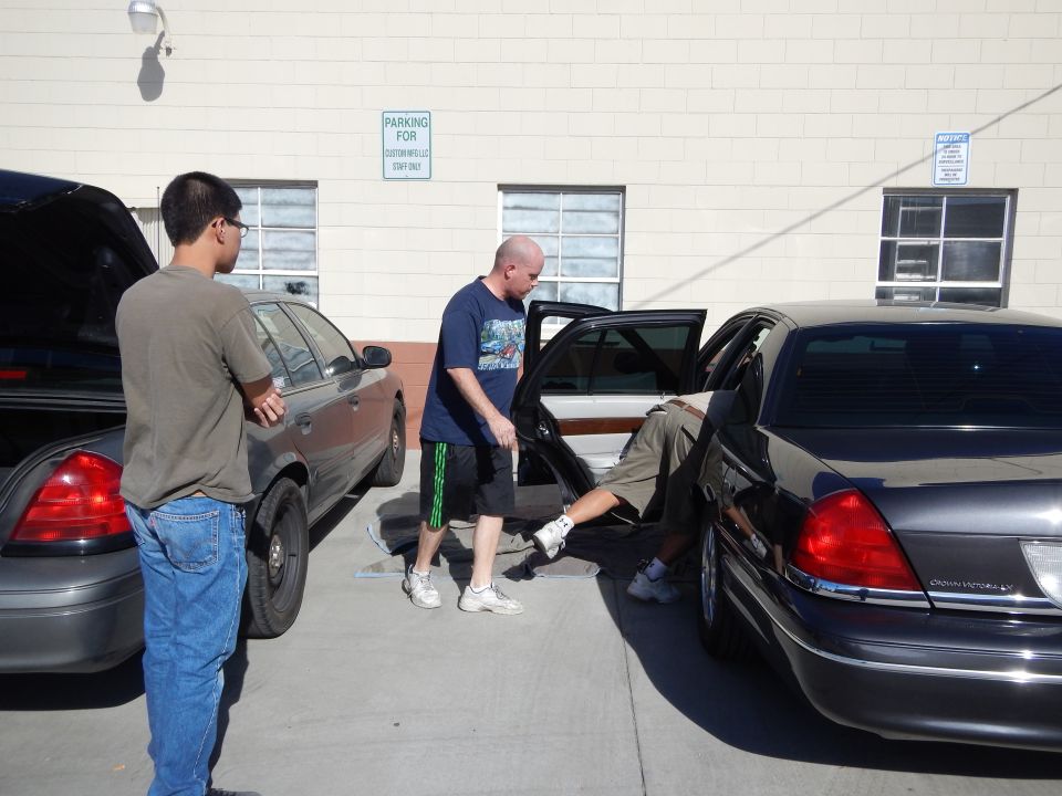 Men working around the open rear door of a dark Lincoln sedan beside a gray Crown Victoria in a lot