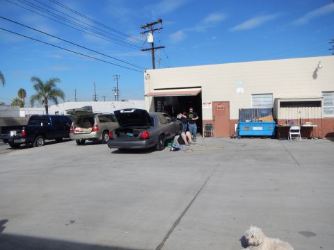 2003 Ford Crown Victoria - wide industrial lot holds a gray Crown Victoria with open trunk, an SUV, a pickup and a dumpster, with people near a roll-up door.