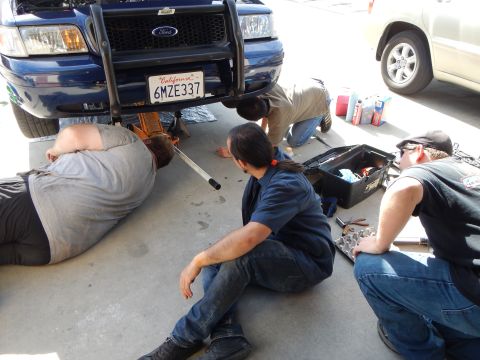 2003 Ford Crown Victoria - Three men work on the ground beneath the front of a blue Ford pickup fitted with a brush guard, California plate 6MZE337.