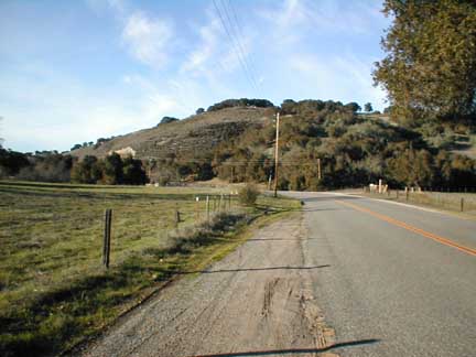 1994 Ford Crown Victoria - A paved road winds through a rural landscape with a grassy field and a distant hill.