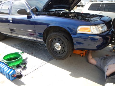 2003 Ford Crown Victoria - blue Ford Crown Victoria is raised on a floor jack with its front steel wheel exposed, oil bottles nearby and a person lying underneath.
