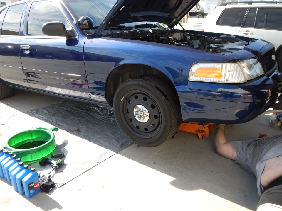 Blue Ford Crown Victoria on a jack with front wheel showing steel rim, oil bottles and a person underneath