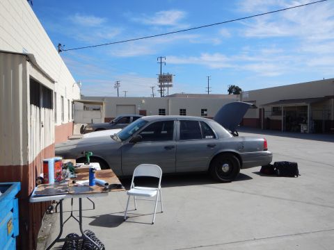 2003 Ford Crown Victoria - silver Ford Crown Victoria sits hood-up beside a white folding chair and a makeshift worktable in a sunny industrial lot.