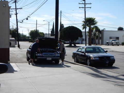 2003 Ford Crown Victoria - Two men stand by a black GMC SUV with its hood raised on a quiet street while a black Crown Victoria sits parked across the road.