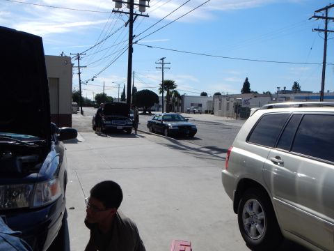 2003 Ford Crown Victoria - tan Toyota SUV and a black sedan share an industrial street lined with power lines and parked cars, with a man in the foreground.