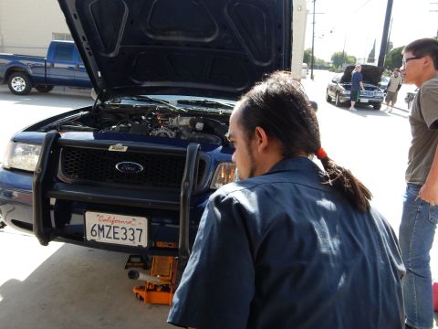 2003 Ford Crown Victoria - ponytailed man inspects the open engine bay of a blue Ford pickup with a brush guard and California plate 6MZE337 in a sunny lot.