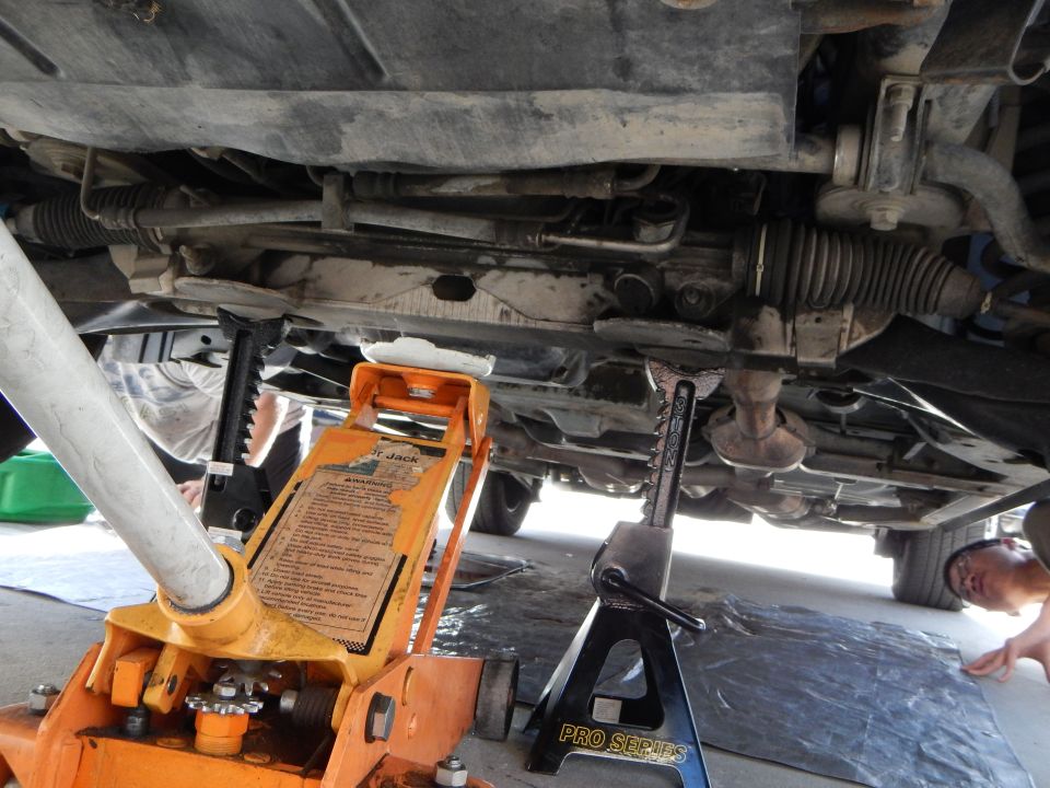 Underbody of a car on an orange floor jack and jack stand showing steering rack and exhaust, person reaching in