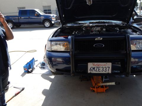 2003 Ford Crown Victoria - blue Ford Crown Victoria with brush guard sits hood-up on a floor jack, with a white Ford Super Duty pickup parked behind it.