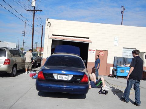 2003 Ford Crown Victoria - blue Ford Crown Victoria sits with its trunk up outside an open roll-up door, plate 6MZE337, while people stand and work nearby.