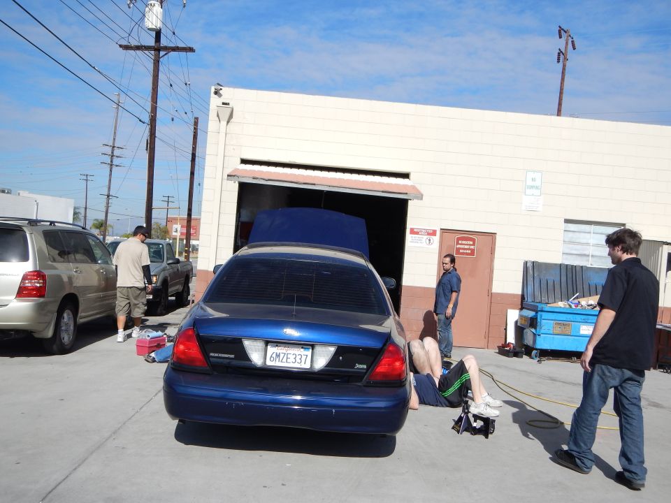 Rear of a blue Ford Crown Victoria with trunk up, plate 6MZE337, outside a roll-up door with people nearby