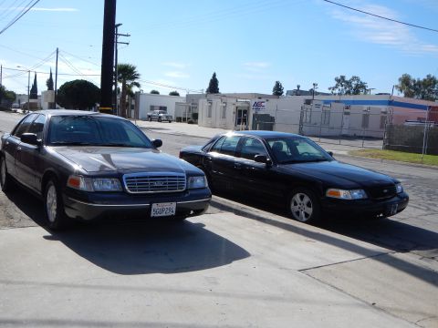 2003 Ford Crown Victoria - gray Ford Crown Victoria with plate 5GUP294 sits beside a black Crown Victoria on a sunny industrial street.