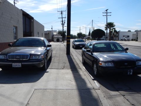2003 Ford Crown Victoria - gray Crown Victoria with plate 5GUP294 and a black Crown Victoria flank a utility pole on a quiet industrial street.