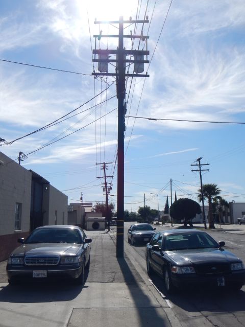 2003 Ford Crown Victoria - tall utility pole topped with transformers rises over an industrial street where gray and black Crown Victoria sedans are parked.