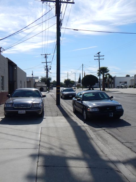 2003 Ford Crown Victoria - Two dark-colored Ford full-size sedans parked on opposite sides of an industrial street under utility poles on a sunny day.