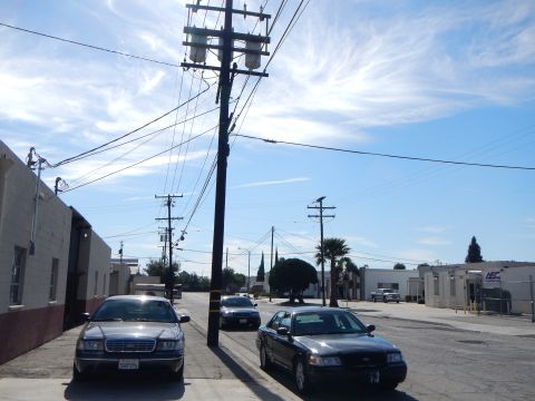 2003 Ford Crown Victoria - Two dark full-size sedans parked along an industrial street lined with single-story commercial buildings and palm trees.