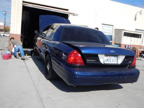 2003 Ford Crown Victoria - Rear three-quarter view of a dark blue Ford Crown Victoria with hood raised outside a garage, California plate 6MZE337.