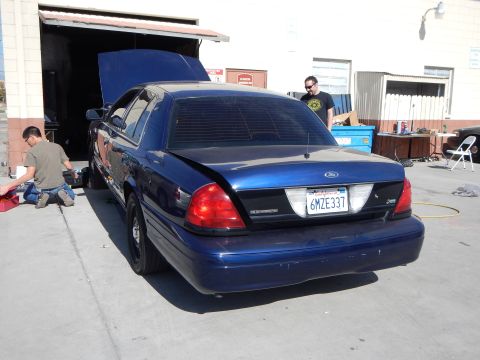 2003 Ford Crown Victoria - Rear of a blue Ford Crown Victoria with raised hood outside a shop; a man stands nearby and another kneels at the front.