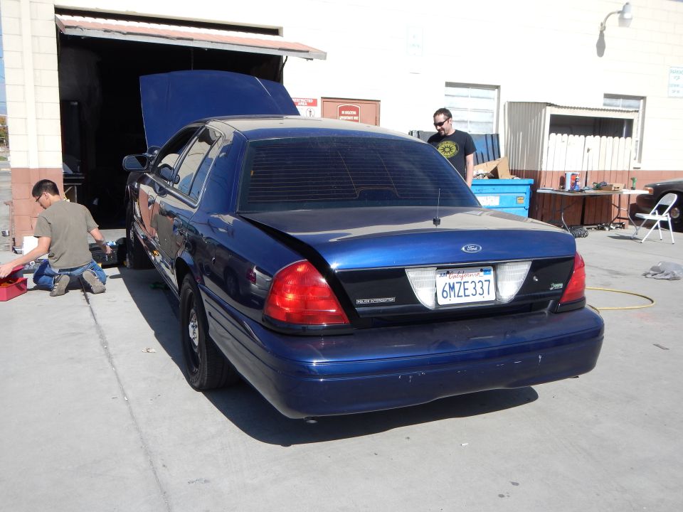 Rear of blue Ford Crown Victoria with open hood, man standing behind, plate 6MZE337