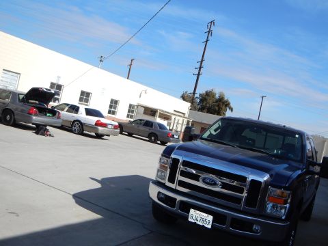 2003 Ford Crown Victoria - dark blue Ford pickup parked in a sunlit lot with silver Ford sedans, one with its trunk open, beside a white building.