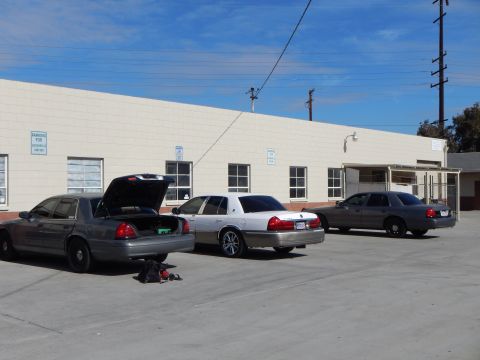 2003 Ford Crown Victoria - Three gray and silver Ford sedans lined up in a lot beside a beige industrial building, the nearest with its hood open.