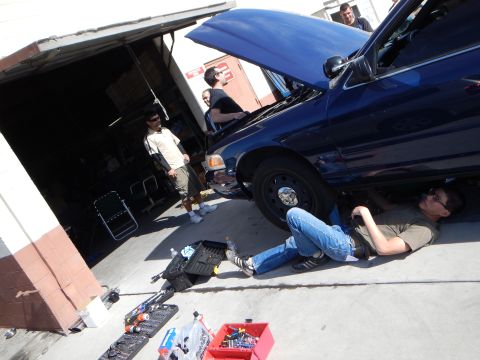 2003 Ford Crown Victoria - Several people work on a dark blue Ford sedan outside a garage, one lying on the pavement under the front with tools spread out.