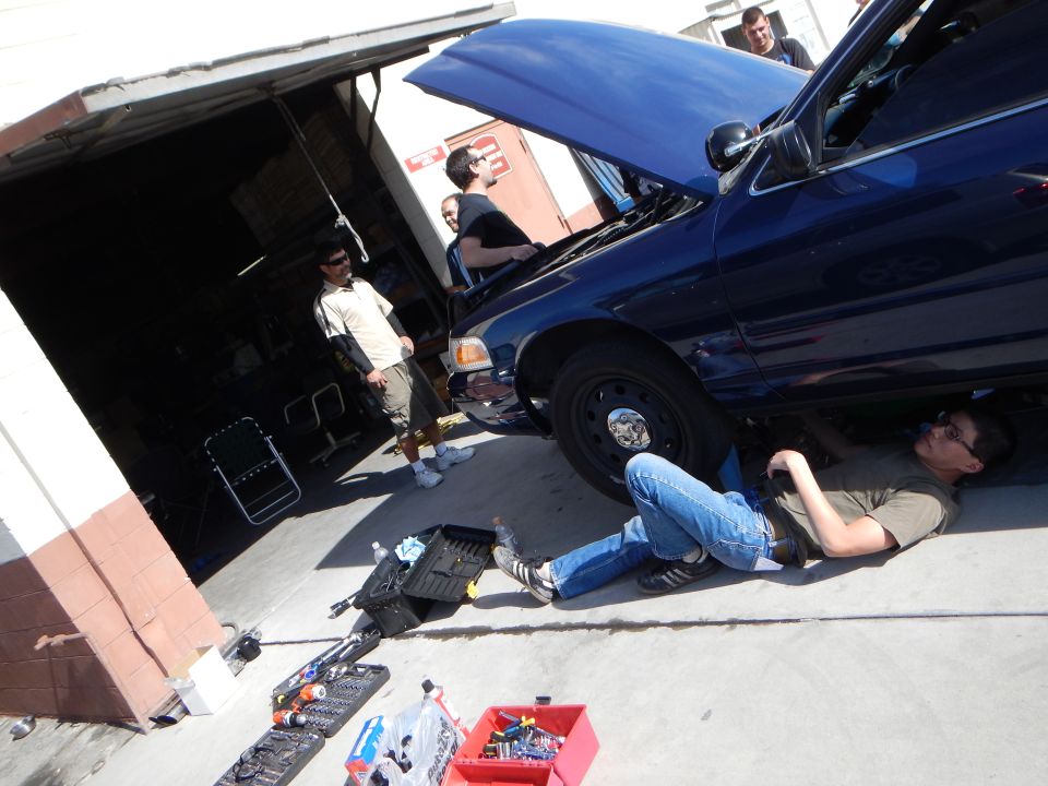 People working on a dark blue Ford sedan, one lying underneath on the pavement