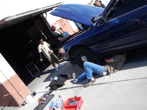 2003 Ford Crown Victoria - group of people work on a dark blue Ford sedan outside a shop, with one man lying under the car amid scattered tools.