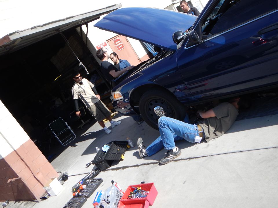 Group working on a dark blue Ford sedan, a man on the ground beneath it