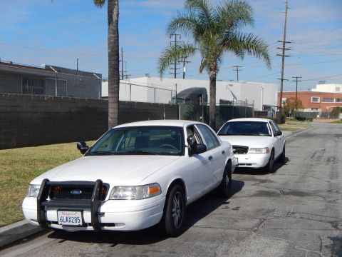 2003 Ford Crown Victoria - white Ford Crown Victoria with a front push bar parked at the curb ahead of a second white sedan, California plate 6LAX285.