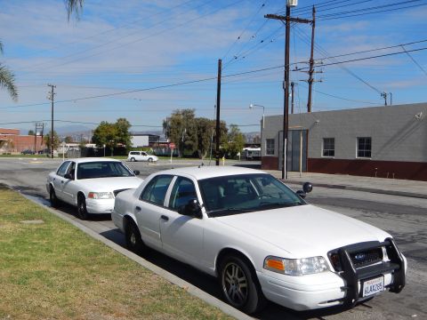 2003 Ford Crown Victoria - Two white Ford full-size sedans, the front one with a push bar, parked at a curb near a gray building under utility lines.