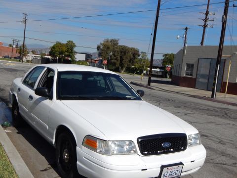 2003 Ford Crown Victoria - white Ford Crown Victoria sedan with black steel wheels parked on a quiet industrial street corner under a blue sky.