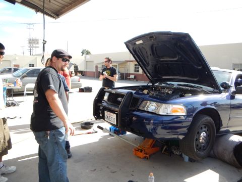 2003 Ford Crown Victoria - Several men stand around a dark blue Ford sedan raised on a jack with its hood open outside an auto shop.