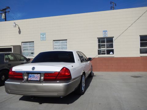 2003 Ford Crown Victoria - Rear three-quarter view of a white-over-tan Mercury Grand Marquis LS sedan parked beside a tan building.