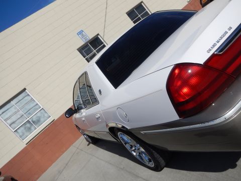 2003 Ford Crown Victoria - Low rear-quarter view of a white-over-tan Mercury Grand Marquis GS sedan with chrome wheels beside a building.