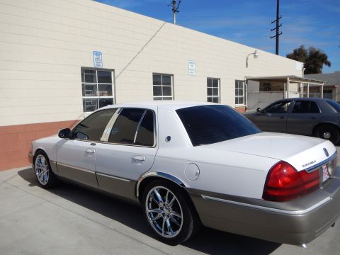 2003 Ford Crown Victoria - Side profile of a white-over-tan Mercury Grand Marquis sedan on chrome wheels with tinted windows parked by a building.