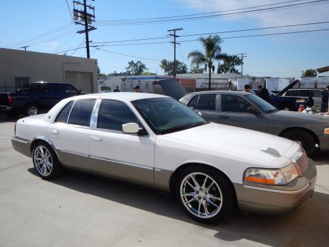 2003 Ford Crown Victoria - Front-quarter view of a white-over-tan Mercury Grand Marquis sedan on chrome wheels in a lot with other cars.