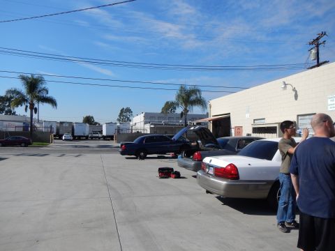 2003 Ford Crown Victoria - Several sedans, including a dark one with its hood up, parked in an industrial lot as two men stand by a white car.