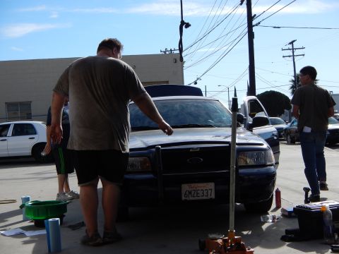 2003 Ford Crown Victoria - man leans over the open hood of a dark blue Ford sedan fitted with a push bar, with tools spread on the ground.