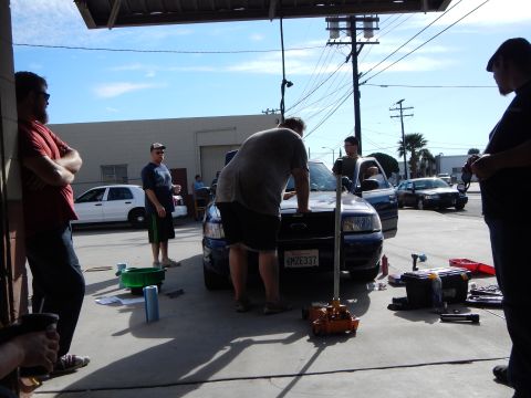 2003 Ford Crown Victoria - Several men gather around a dark blue Ford sedan with a push bar being worked on outside a shop, tools on the ground.