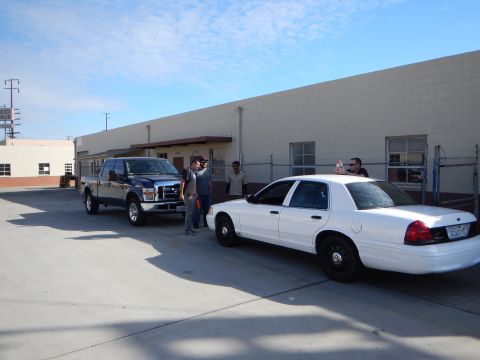 2003 Ford Crown Victoria - dark Ford crew-cab pickup truck and a white Ford Crown Victoria sedan parked beside a building as people stand nearby.