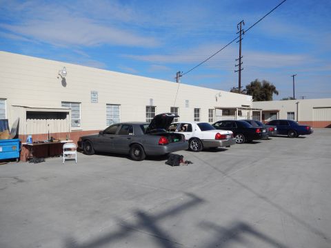 2003 Ford Crown Victoria - row of gray, white, and dark sedans parked along a long building in an industrial lot under a blue sky.
