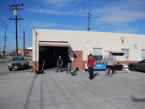 2003 Ford Crown Victoria - Several people stand in a paved lot outside the open roll-up door of a beige industrial garage building under a clear blue sky.