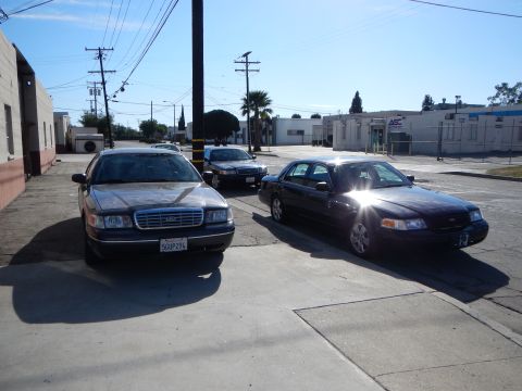 2003 Ford Crown Victoria - Three dark full-size Ford sedans, one with a visible Ford grille emblem, parked at angles in a sunny industrial lot.