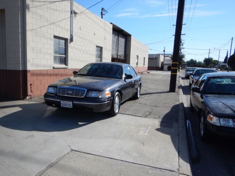 2003 Ford Crown Victoria - dark gray Ford sedan with a front license plate sits angled beside a cinderblock industrial building, with more cars lining the curb.