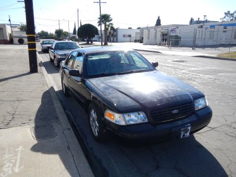 2003 Ford Crown Victoria - black Ford sedan with the Ford grille emblem is parked along a sunny curb, with several similar sedans queued behind it.