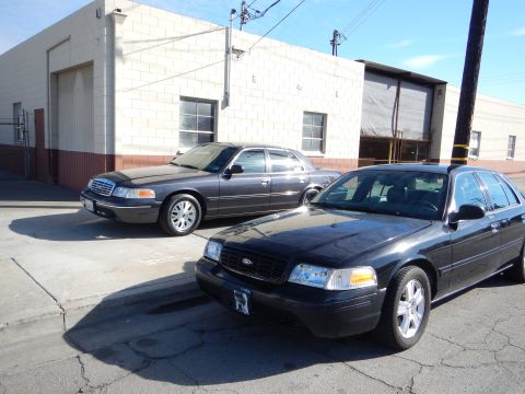 2003 Ford Crown Victoria - Two dark Ford sedans, both showing the Ford grille emblem, sit parked on cracked pavement outside a cinderblock industrial building.