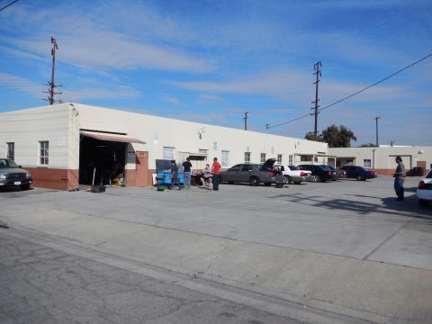 2003 Ford Crown Victoria - long beige industrial warehouse with an open garage door, several parked cars and people working in the lot under a blue sky.
